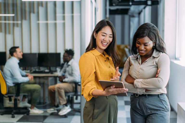 Two women stand in an office hallway, engrossed in a tablet that showcases financial workflow automation. One wears a yellow blouse, the other a light gray one. Behind them, two people are seated at desks, illustrating the seamless integration of technology in their work environment.