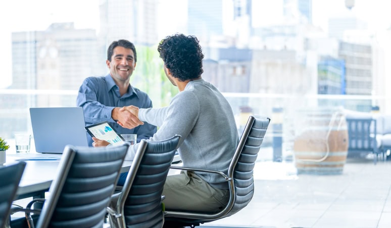 Two men shake hands across a conference table in a modern office with large windows and city views. One is holding a tablet displaying charts.