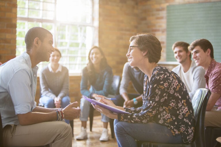 A group of people sit in a circle during a discussion in a bright room with a large window and brick walls.