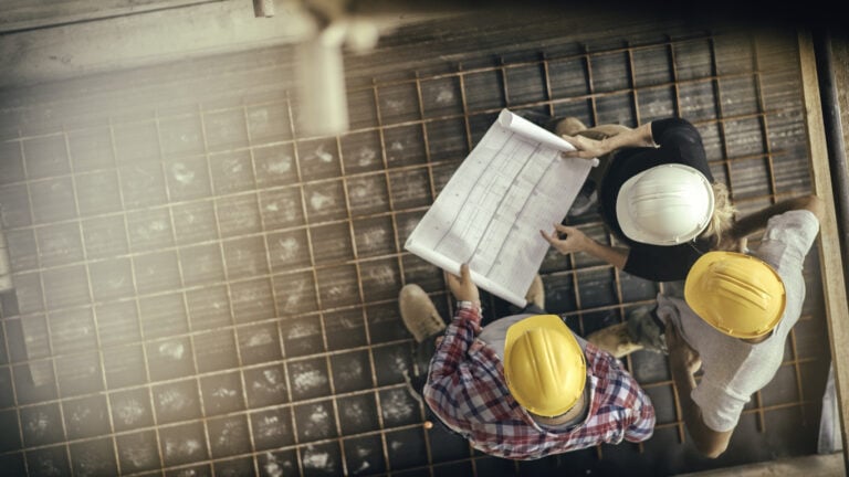 Overhead view of three construction workers with hard hats examining a blueprint on a building site.