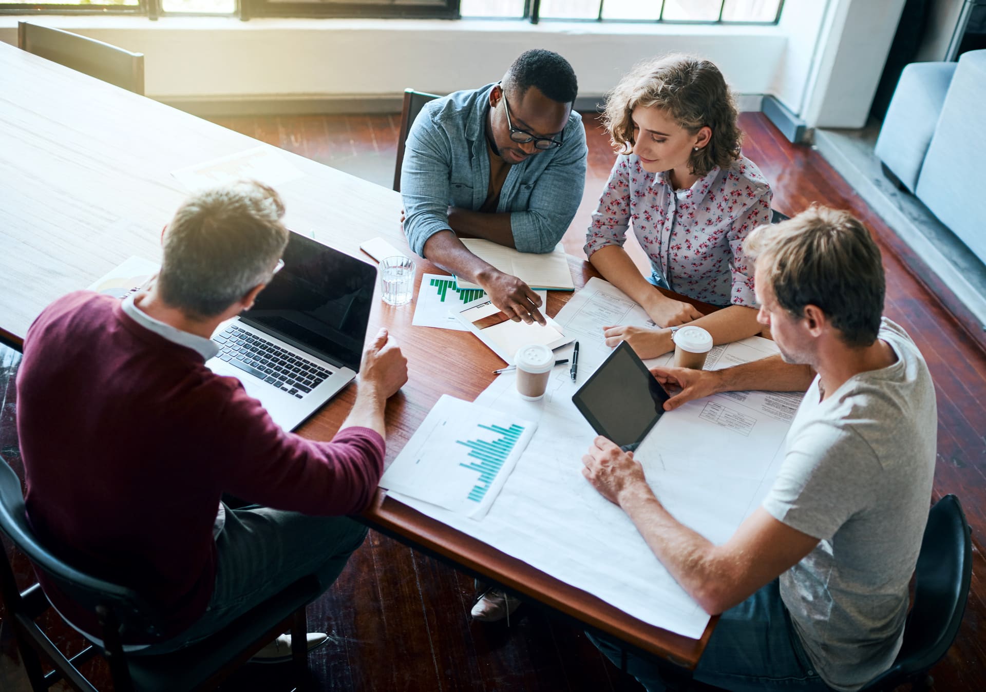 Four people are gathered around a table with documents, charts, and laptops, engaged in a discussion.