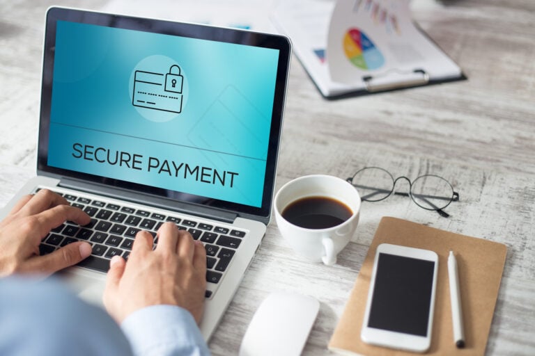 SECURE PAYMENT CONCEPT Person typing on a laptop displaying "Secure Payment" with a lock icon, beside a coffee cup, glasses, notebook, and pen on a desk.