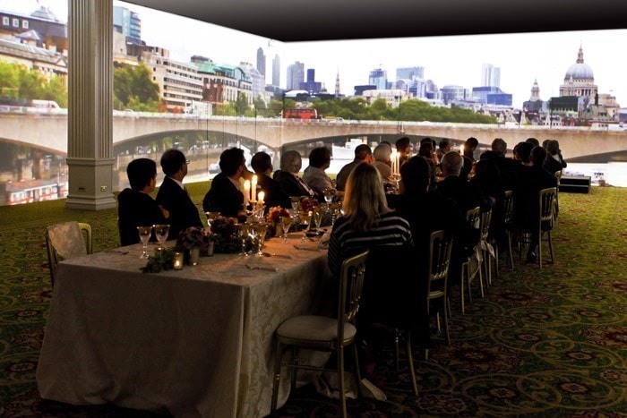 A group of people sit at a long dining table in a room with large projector screens displaying a cityscape with a bridge and skyline, creating an immersive dining experience.