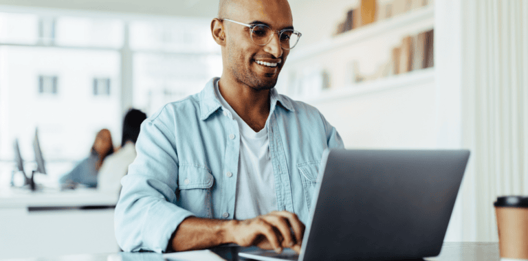 A person wearing glasses and a denim shirt is smiling while managing business expenses on a laptop in a bright room. Shelves with books are in the background.