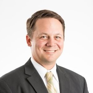 A man in a dark suit and light tie smiles at the camera against a plain white background.