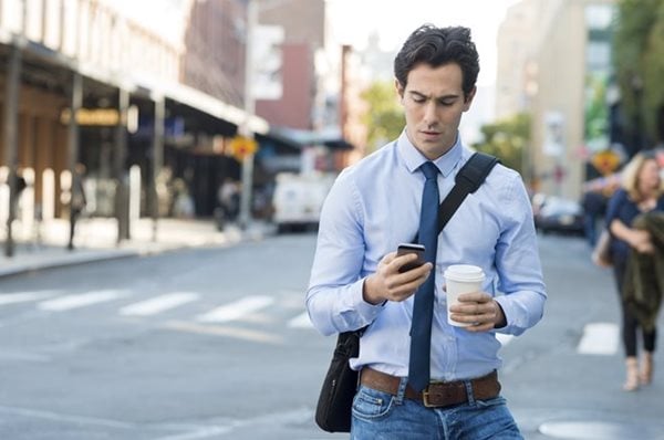 A man in a blue shirt and tie walks on a city street, holding a smartphone and a coffee cup. He has a bag slung across his shoulder.