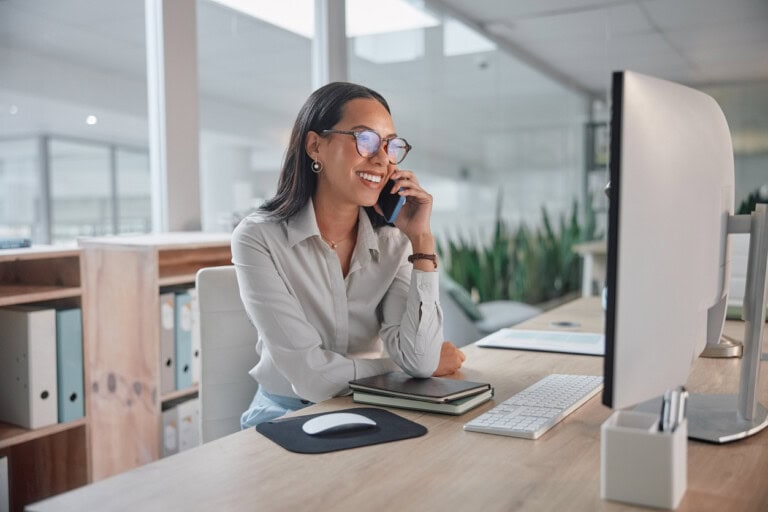 A woman sits at a desk in a modern office, smiling while talking on a mobile phone, with a computer monitor, keyboard, and notebooks in front of her.