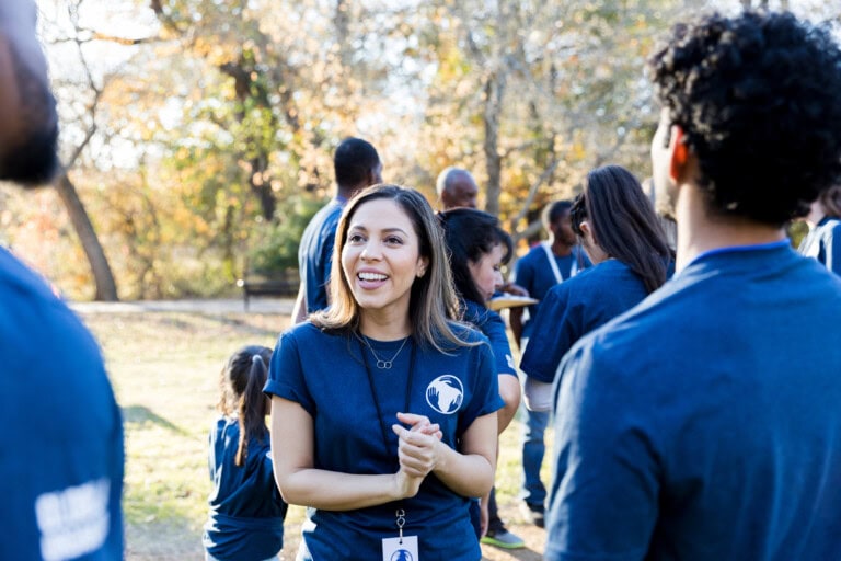 A group of people wearing matching blue shirts are gathered outdoors, talking about their nonprofit expenses and smiling, with trees and sunlight in the background.