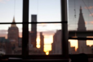 nyc_sunrise-300x2001.jpg Blurred city skyline at sunset viewed through window with partially closed blinds.
