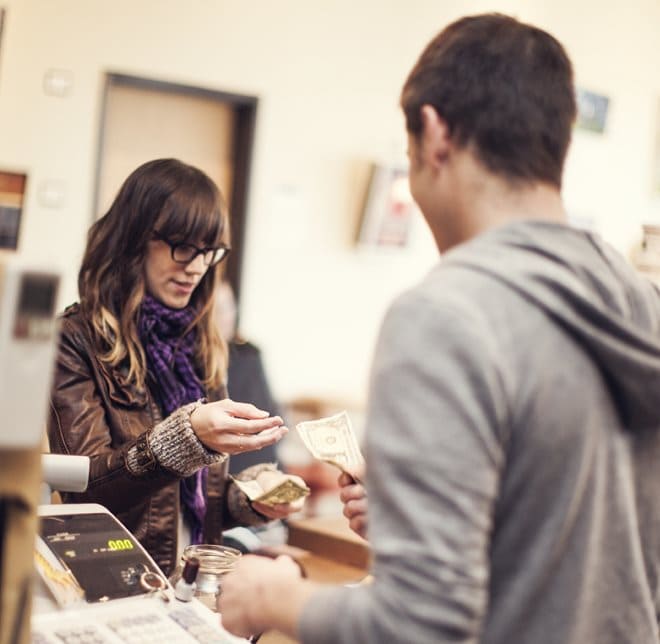 pettycash2.jpg A woman in a brown jacket and scarf gives change to a man in a gray hoodie at a checkout counter.