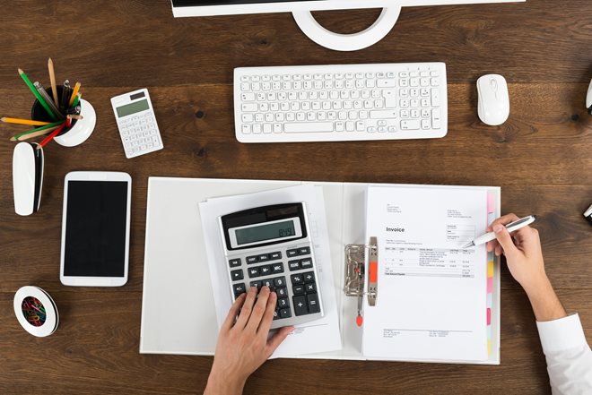 Person working at a desk with a calculator, keyboard, tablet, and invoices.