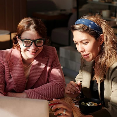 Two people sit at a table looking at a laptop, with food bowls in front of them. One has a headband, the other wears glasses. Sunlight streams in, creating a warm atmosphere.