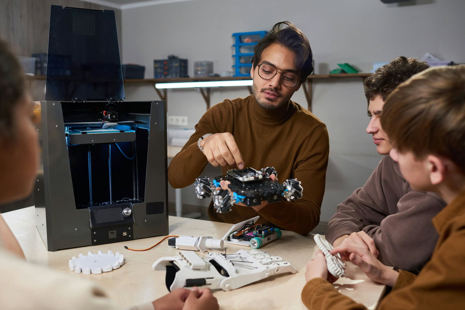 school-robotics-program A man demonstrates a robotic device to three people at a table; a 3D printer and various components are visible.