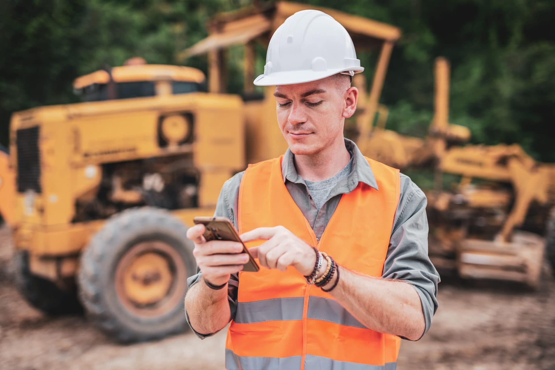 A construction worker in an orange vest and white hard hat uses a smartphone at a construction site with heavy machinery in the background.