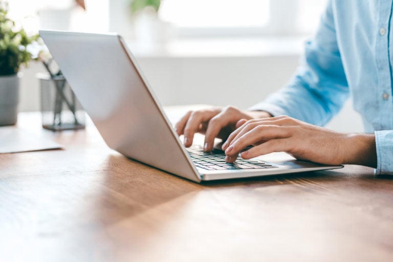 Person typing on a laptop at a wooden table, with a plant in the background.