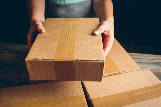 A person holding a brown cardboard box with packing tape on a wooden surface, surrounded by three similar boxes.