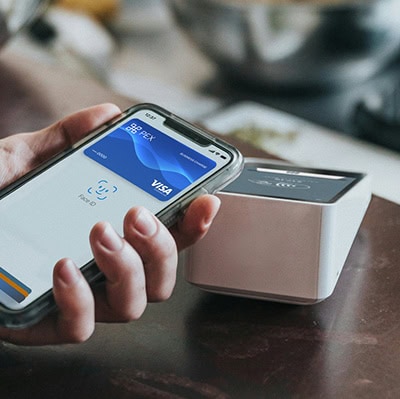 A hand holding a smartphone with a digital Visa card displayed, making a contactless payment over a point-of-sale terminal.