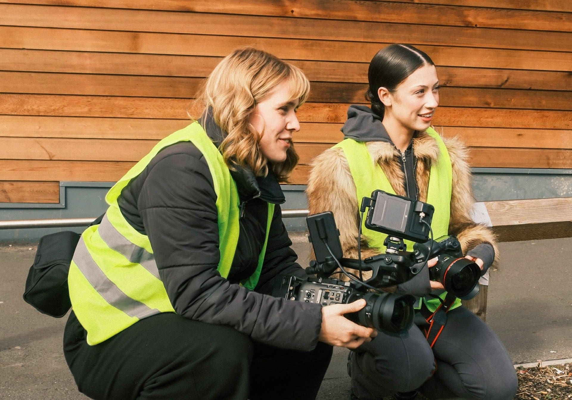 team-filming Two people crouch outside, wearing high-visibility vests and holding camera equipment.