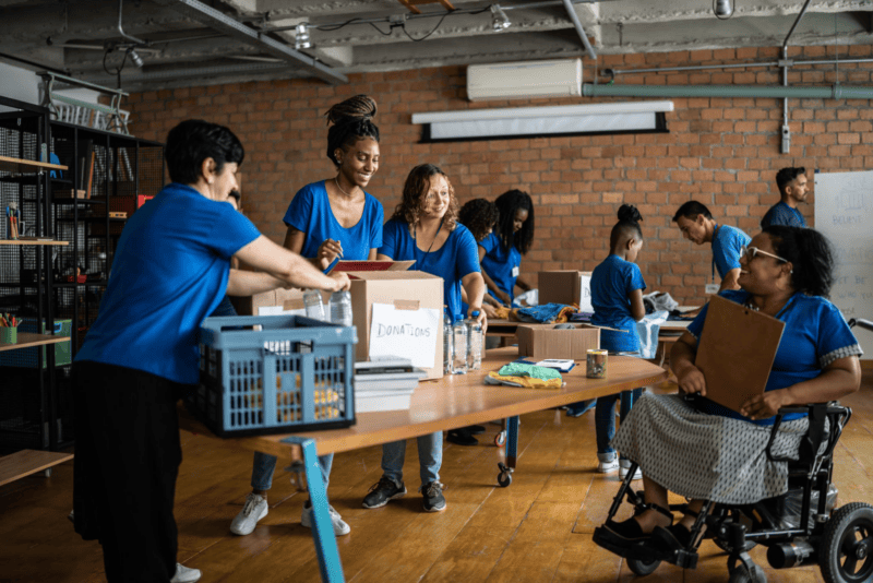 volunteer-donation-center A group of volunteers in blue shirts organize donation items on tables in a community center. A woman in a wheelchair participates.