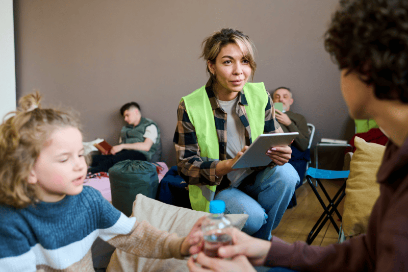 volunteer-worker A woman in a reflective vest holds a tablet while talking to a seated person. A child and another adult sit nearby. Background shows people resting on cots.