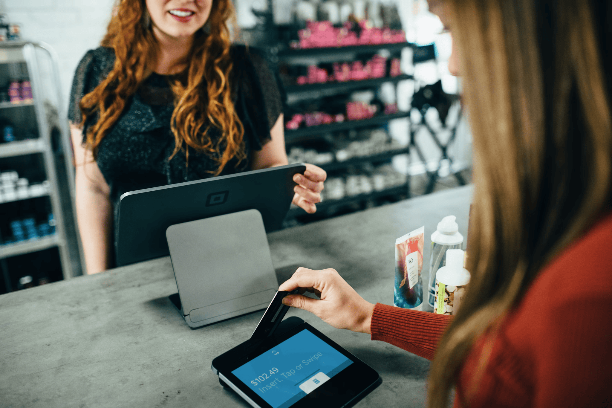 woman-paying-at-salon A person is using a credit card to pay on a tablet device at a counter, while a cashier stands nearby.